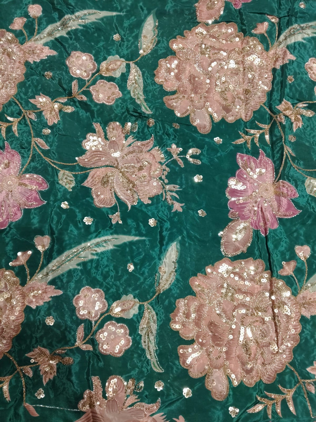A close-up of a teal green brocade fabric featuring large, detailed floral motifs in pink and rose gold, embellished with shimmering sequins and metallic embroidery.