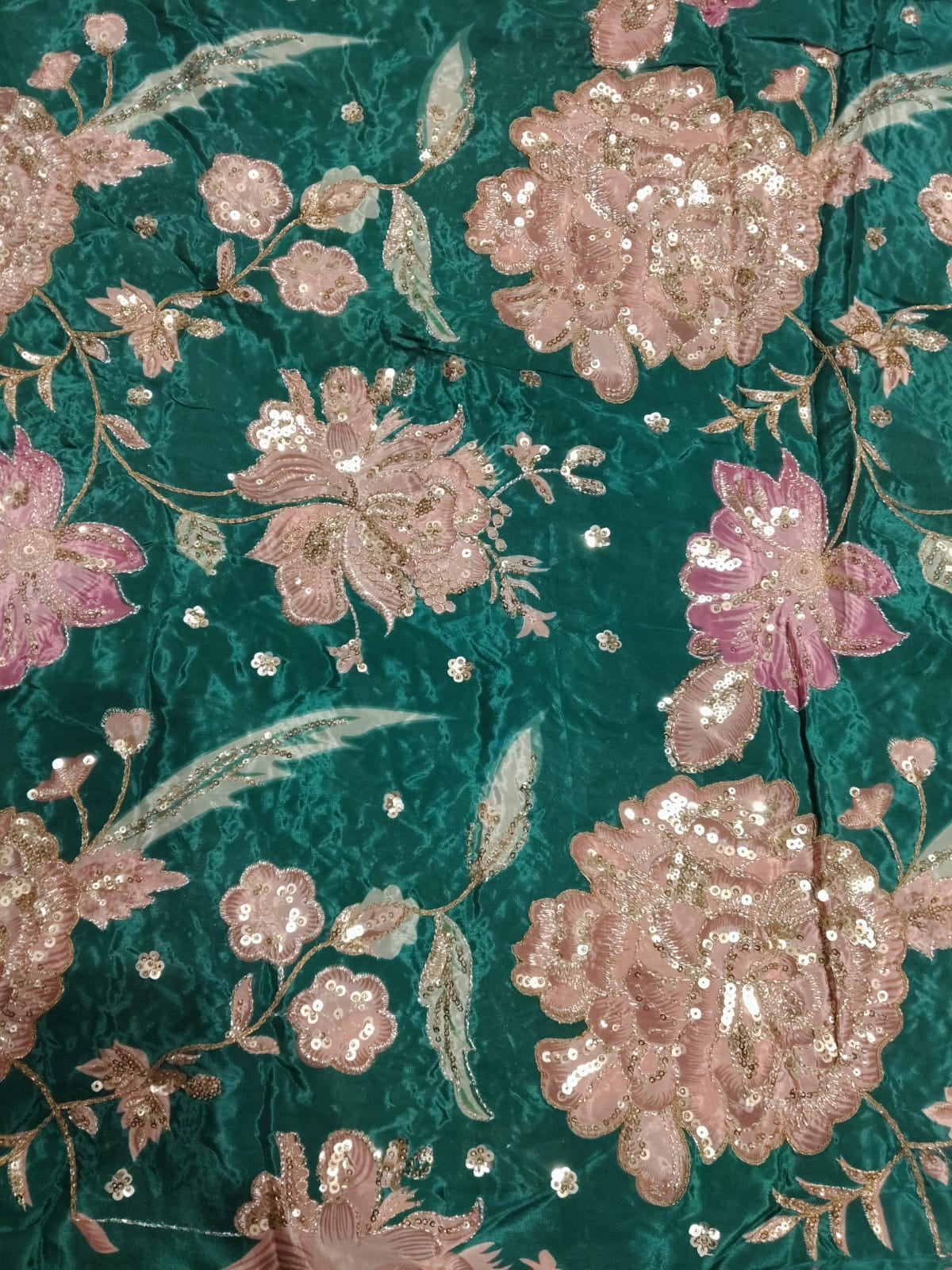 A close-up of a teal green brocade fabric featuring large, detailed floral motifs in pink and rose gold, embellished with shimmering sequins and metallic embroidery.