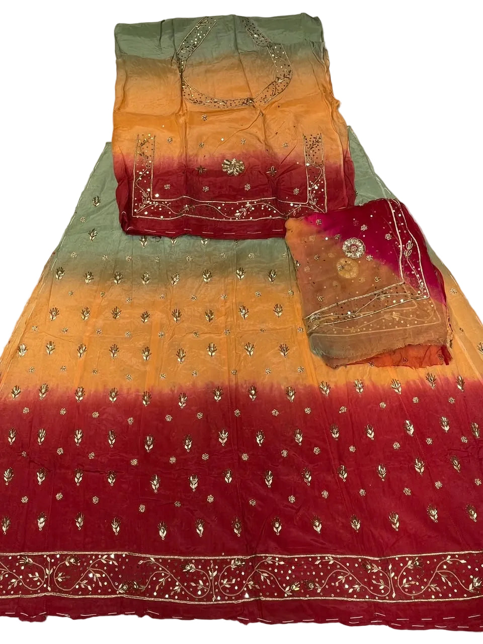 A folded and spread-out traditional Indian ethnic outfit, likely a salwar suit set, featuring a vibrant orange, yellow, and red gradient fabric with silver mirror work embroidery and a contrasting red dupatta.