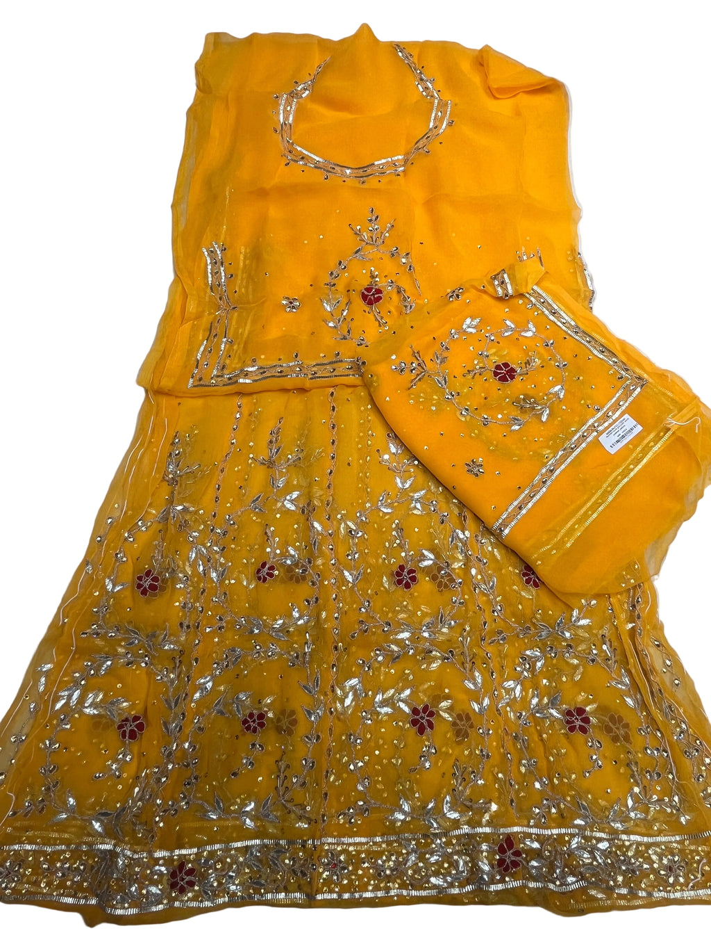 A bright yellow traditional embroidered outfit laid flat on a white sheet, including a kurta or dress, matching pants, and a small pouch or clutch. The fabric is heavily decorated with silver mirror work and floral embroidery with small red accents.