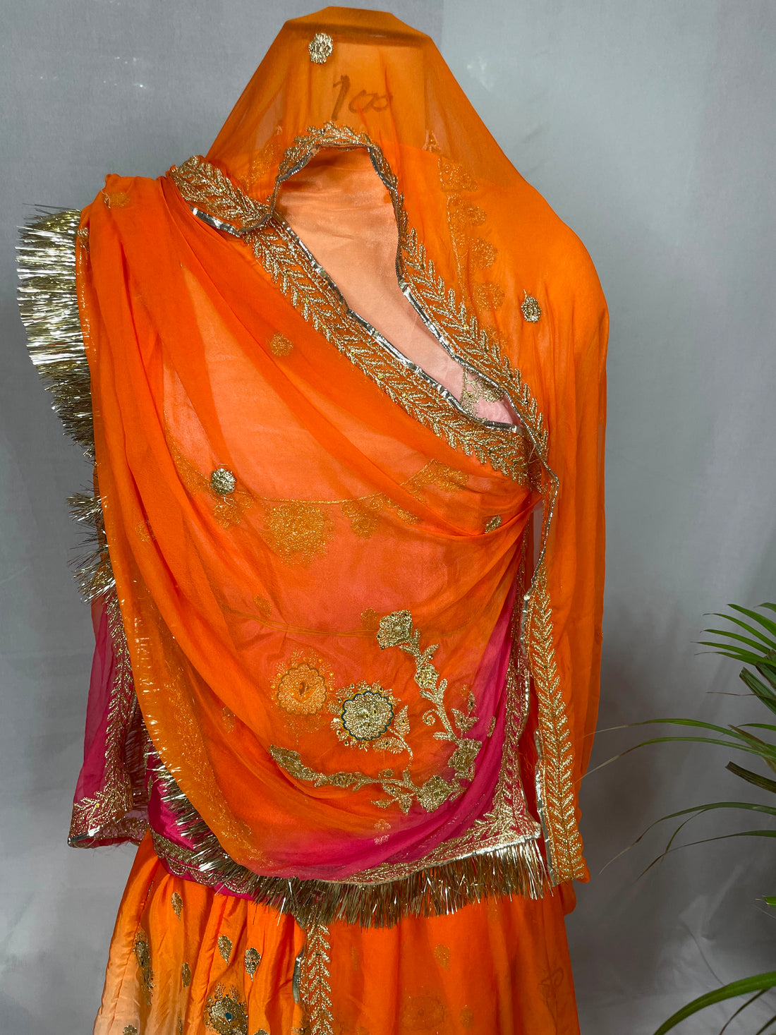 An orange and pink traditional draped garment with gold embroidery and metallic fringe, displayed on a mannequin against a gray wall with a plant leaf at the side.