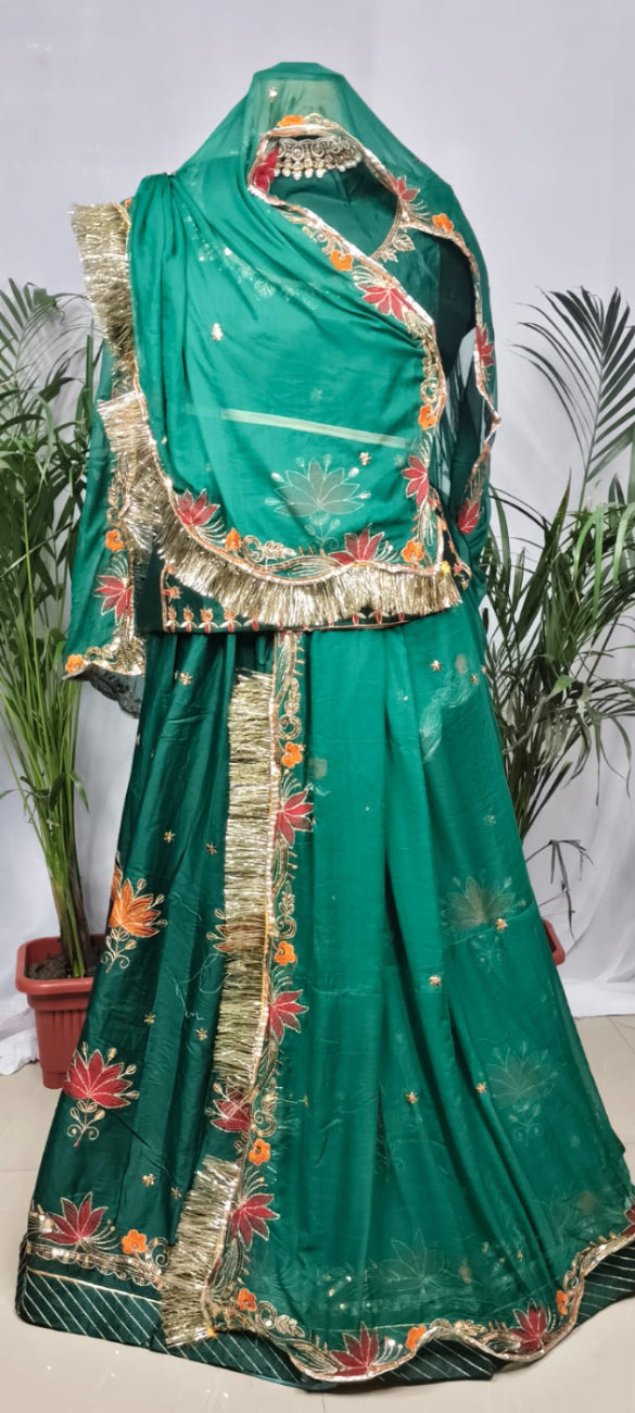 A green traditional outfit displayed on a mannequin, featuring a flared skirt, matching draped fabric, and ornate gold embroidered borders with red floral accents, set against a white backdrop with potted plants.