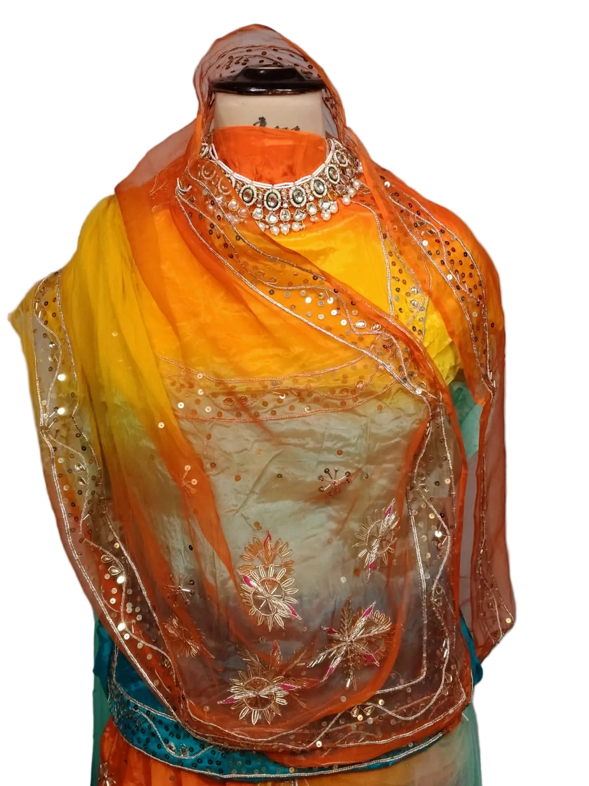 A traditional Indian outfit displayed on a mannequin, featuring a yellow and orange gradient dupatta with silver mirror work, a yellow and orange printed blouse with a silver mirror-studded collar, and a teal and orange skirt.