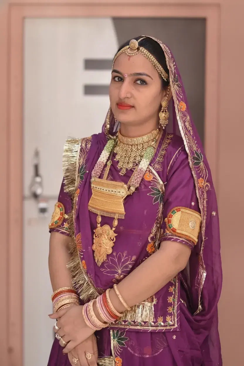 A woman wearing an ornate purple traditional outfit with gold embroidery, a matching head covering, heavy gold jewelry, and stacked bangles, standing indoors in front of a soft pink door.